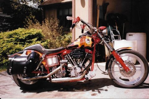 1989 Ford Bronco - A custom red and flame-painted motorcycle with a black leather saddlebag and helmet is parked on a concrete driveway in front of a garage.