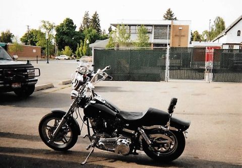 1989 Ford Bronco - A black motorcycle is parked on asphalt in front of a chain-link fence and a modern building. A dark-colored SUV is partially visible on th…