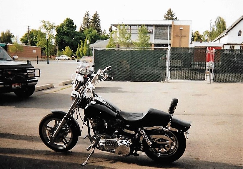 Black motorcycle parked in front of a fence and building, with a dark SUV to the left.