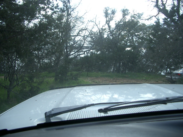 White vehicle hood and windshield wipers, with trees and a distant body of water visible through the windshield