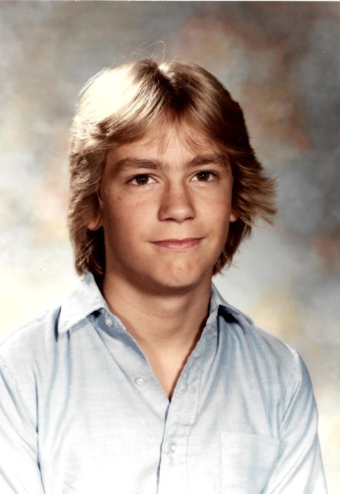 1989 Ford Bronco - A headshot of a young boy with feathered blonde hair, wearing a light blue collared shirt. The background is a mottled grey and beige studi…