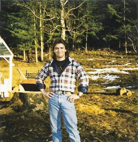1989 Ford Bronco - A man stands in a wooded area with patchy snow on the ground. He is wearing a plaid shirt and jeans.