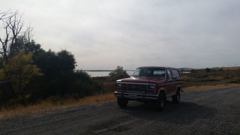 1981 Ford Bronco - A red 1980s Ford Bronco is parked on a gravel road next to a body of water and dense vegetation. The vehicle is viewed from a 3/4 front ang…