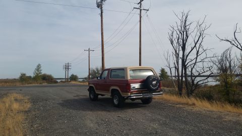 1981 Ford Bronco - A red and tan Ford Bronco is parked on a gravel road next to a body of water. The vehicle is viewed from the rear quarter angle, with power…