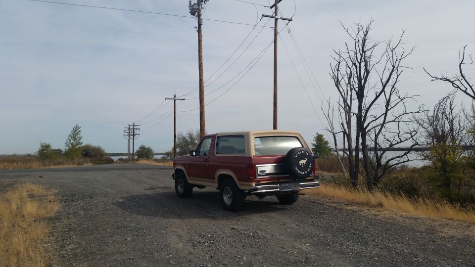 Red and tan Ford Bronco parked on a gravel road near water, with power lines and trees.