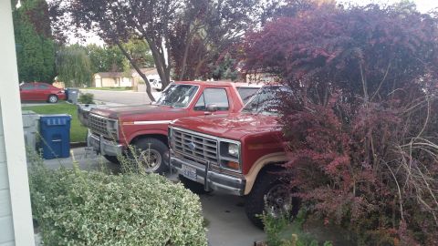 1981 Ford Bronco - Two red Ford trucks are parked next to each other in a driveway. The truck in the foreground has a chrome bumper and a tan stripe along the…