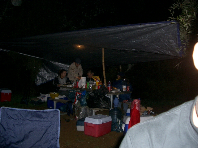 People sit at tables under a large tarp at night, with coolers and camping gear around