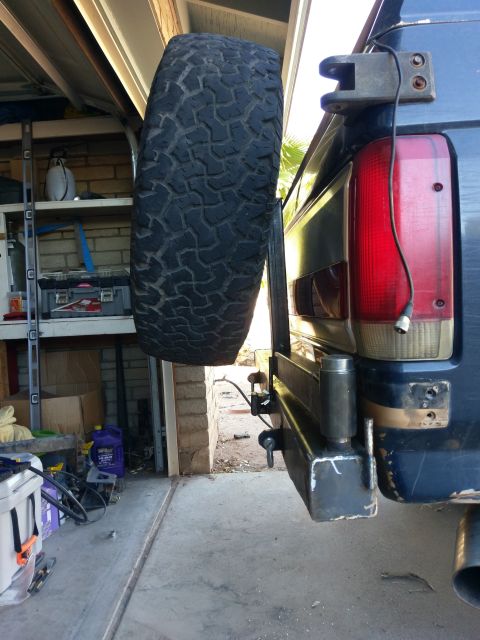 1988 Ford Bronco - A rear-side view of a dark blue SUV with a spare tire mounted on a swing-away carrier attached to a custom rear bumper. The vehicle is park…