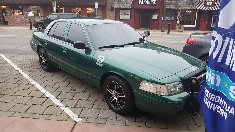 Green Ford Crown Victoria with push bar and number 12 on fender, parked on brick sidewalk.