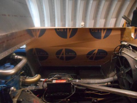 1992 Ford Bronco - The engine bay of a vehicle, showing a blue engine block, exhaust pipes, a battery, and a fan. A large piece of cardboard with blue circula…