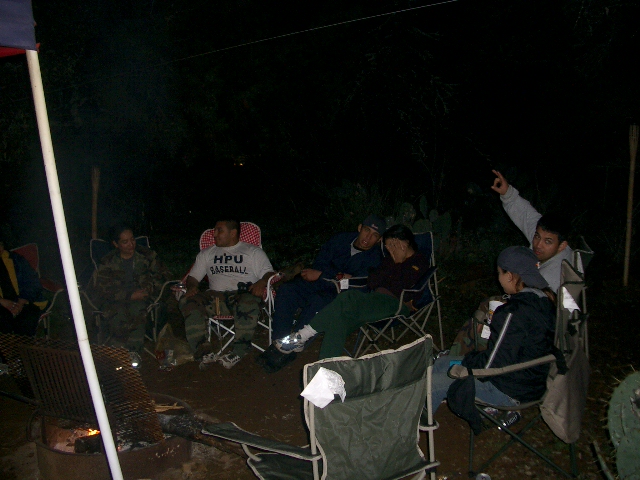 Group of people sitting in camping chairs around a fire pit at night. One person points into the darkness