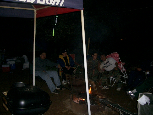 People sitting around a campfire under a tent at night. One person wears a shirt that says HPU BASEBALL