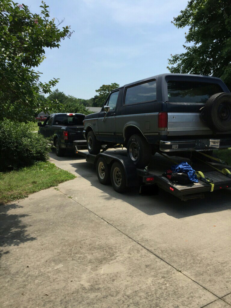 1987 Ford Bronco on a trailer, being towed by a black pickup truck