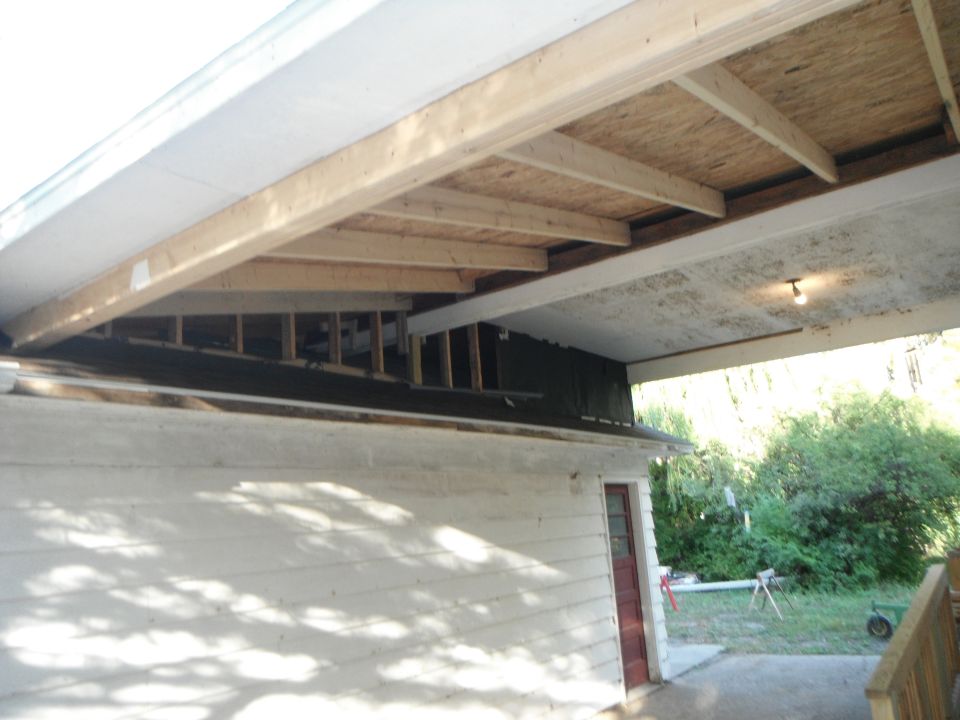 Construction scene showing exposed roof framing and siding of a building, with trees and grass in the background