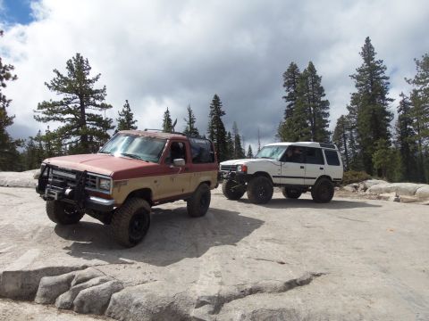 1987 Ford Bronco II - Two modified SUVs navigate a rocky, off-road terrain under a cloudy sky.