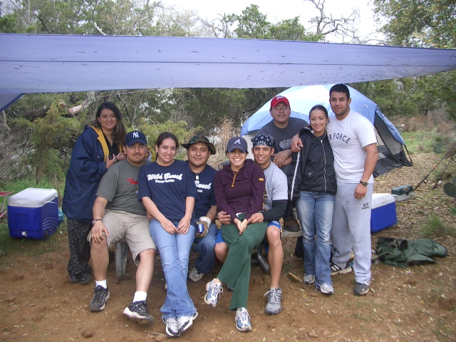 Group of people sitting under a tarp with a tent in the background