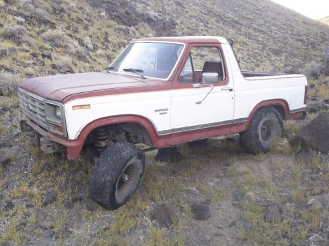 1984 Ford Bronco - A red and white Ford Bronco is parked on a rocky, grassy hillside. The vehicle is angled towards the viewer's left, showing its front and s…