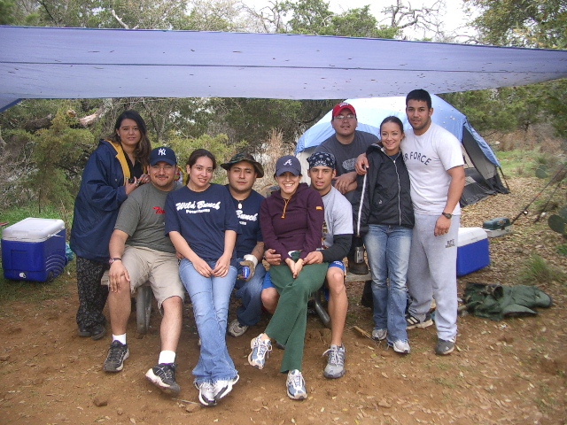 Group of people sitting under a tarp at a campsite with tents and coolers