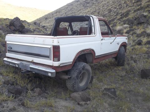 1984 Ford Bronco - The rear driver's side quarter view of a white and red Ford Bronco is parked on a rocky, grassy hillside. The vehicle has a removable soft…