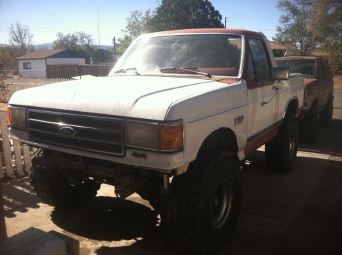 1984 Ford Bronco - A lifted, two-tone white and red Ford pickup truck is parked outdoors in front of another red vehicle. The truck has large off-road tires a…