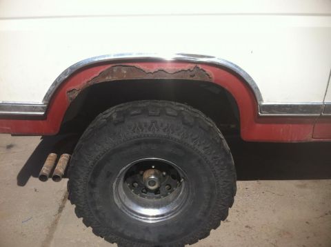1984 Ford Bronco - A close-up side view of the rear wheel well of a white and red Ford Bronco shows significant rust on the fender flare. The vehicle has larg…