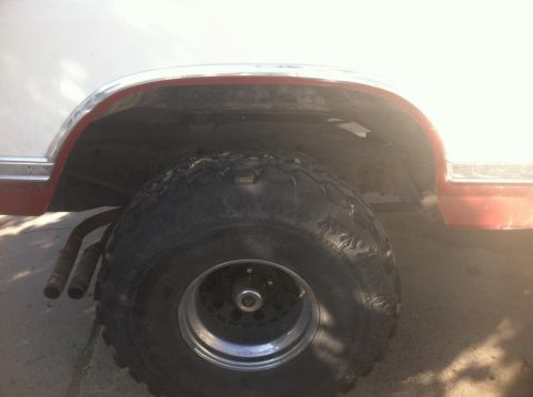 1984 Ford Bronco - A close-up side view of the rear wheel well of a white and red vehicle, likely a 1984 Ford Bronco, showing a large off-road tire and dual e…