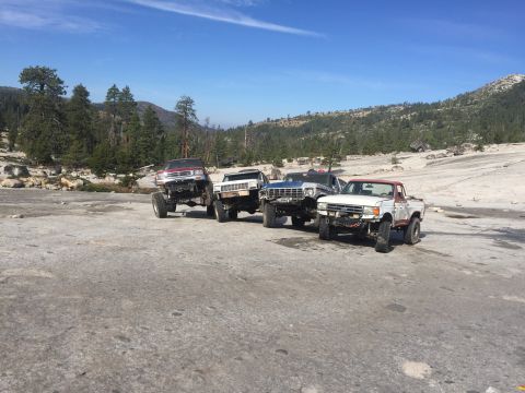 1984 Ford Bronco - Four modified off-road trucks are parked on a rocky, uneven surface with a wooded hillside and blue sky in the background. The trucks are p…