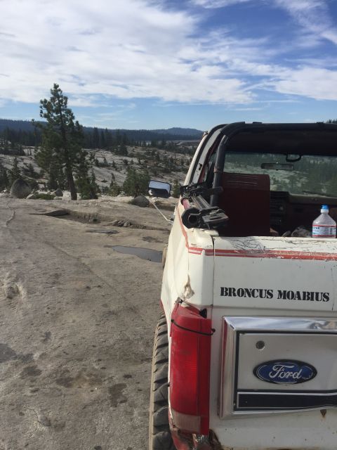 1984 Ford Bronco - The rear passenger side of a white Ford Bronco with a convertible top is parked on a rocky, uneven surface with sparse trees and hills in t…