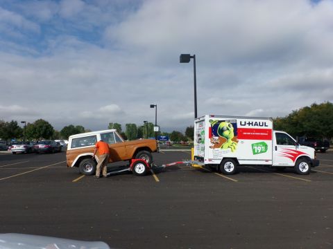 1969 Ford Bronco - A U-Haul truck tows an orange Ford Bronco, likely a first-generation model, across a parking lot.