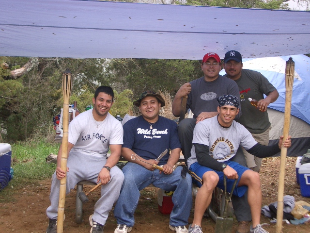 Five men pose for a photo under a tarp at a campsite. One man wears an 'Air Force' shirt, another a 'Wild Bunch' shirt
