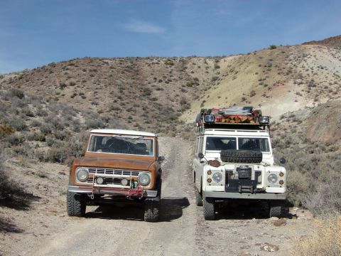 1969 Ford Bronco - A rusty brown 1969 Ford Bronco and a white Land Rover Series III are parked on a dirt road in a desert landscape.