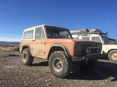1969 Ford Bronco - A muddy, orange 1969 Ford Bronco is parked on a gravel lot next to a white Land Rover with a roof rack. The Bronco is seen from a 3/4 front…