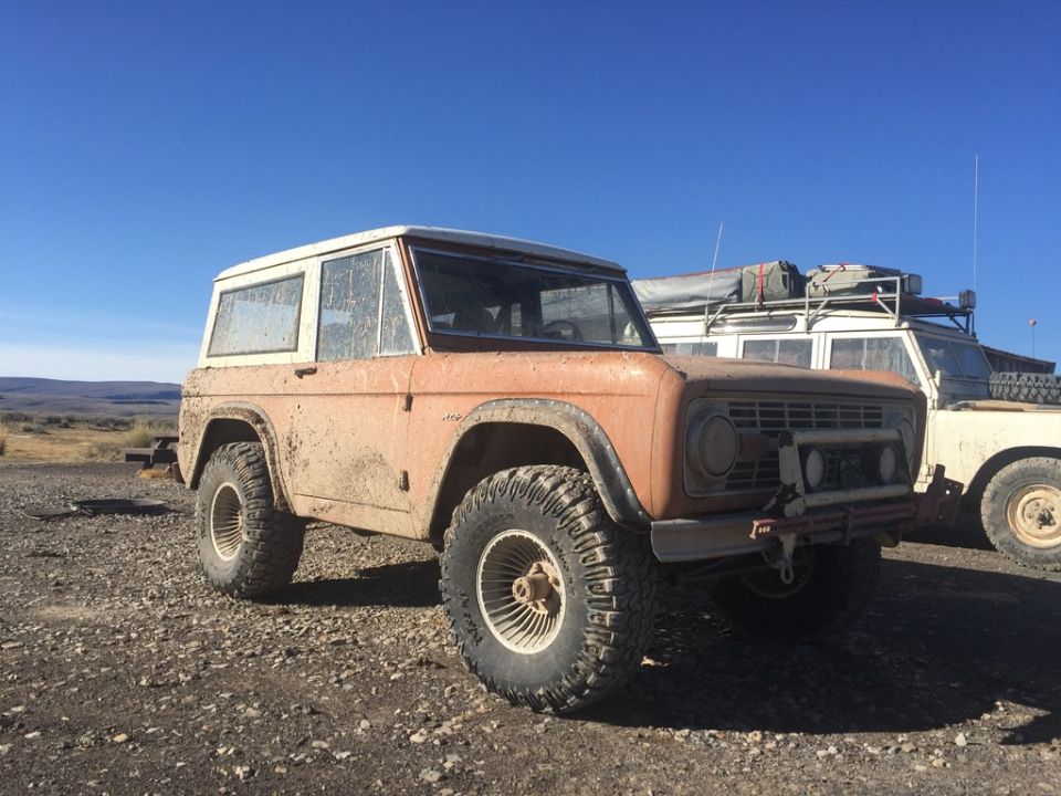 Muddy orange 1969 Ford Bronco parked on gravel next to a white Land Rover.