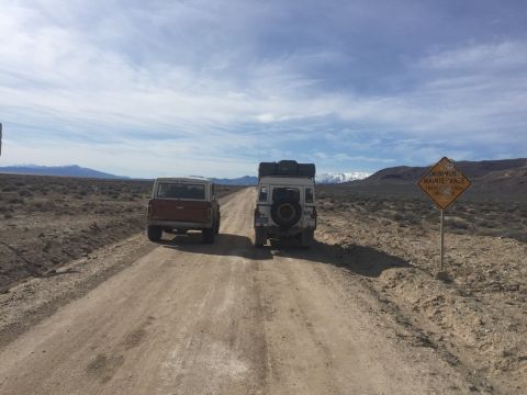 1969 Ford Bronco - Two off-road vehicles are parked on a dirt road in a desert landscape. A diamond-shaped yellow sign with black text is visible to the right…