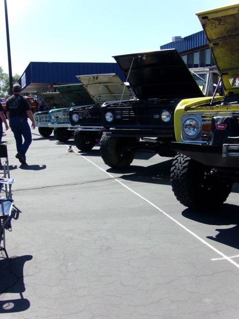 1969 Ford Bronco - A line of Ford Broncos with their hoods open are parked on asphalt under a clear blue sky. The Broncos are various colors including yellow,…