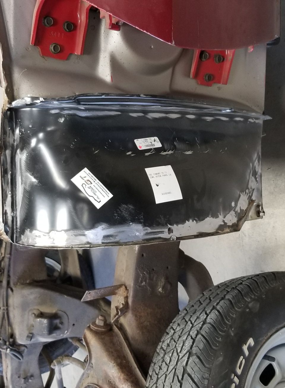 Newly installed black metal cowl outer panel with labels, above rusted car suspension and tire.