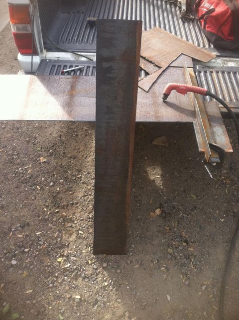 1984 Ford Bronco - A tall, dark metal beam stands vertically in the foreground, casting a shadow on the gravel and dirt ground. Behind it, the tailgate of a w…