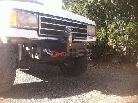 1984 Ford Bronco - The front end of a white Ford truck or SUV is shown, featuring a custom black bumper with a winch and red tow chain. The vehicle is parked…