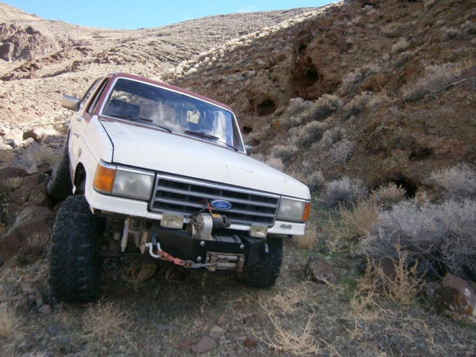 White and red 1980s Ford Bronco with a front winch on a rocky hillside.