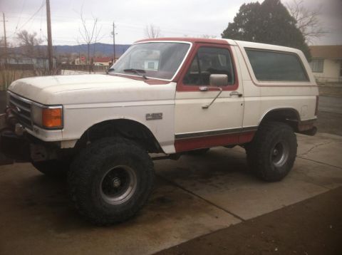 1984 Ford Bronco - A lifted, two-tone white and red 1984 Ford Bronco is parked on a concrete driveway under an overcast sky. The vehicle features large off-ro…