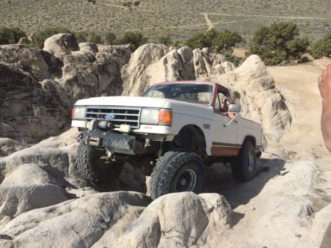 1984 Ford Bronco - A white and red Ford pickup truck is navigating a rocky, off-road trail. The truck has large tires, a winch on the front bumper, and appear…