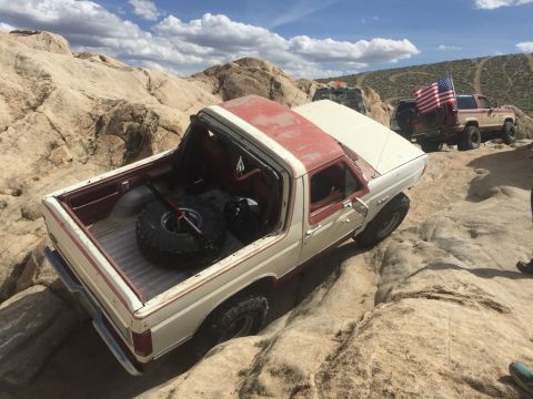 1984 Ford Bronco - A white and red 1980s Ford Bronco is navigating a rocky, off-road trail. The spare tire is visible in the open bed of the truck.