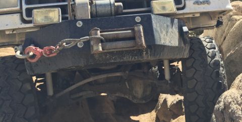 1984 Ford Bronco - The front bumper and winch of an off-road vehicle are shown, with a red tow shackle attached to a cable. The vehicle's large, knobby tires…