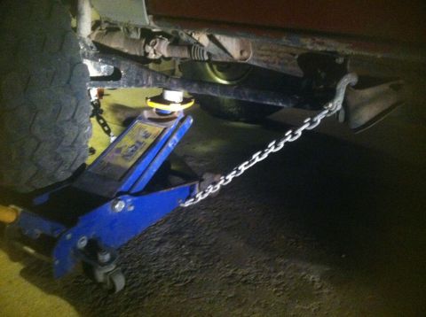 1984 Ford Bronco - A blue hydraulic floor jack is positioned under the frame of a vehicle, supporting it. A metal chain is attached to the vehicle's frame and…