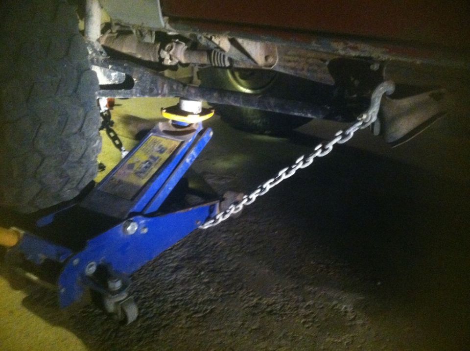 Blue hydraulic floor jack supporting a vehicle frame with a chain attached.