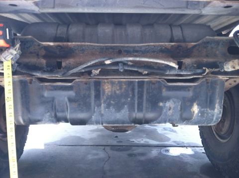 1984 Ford Bronco - The underside of a vehicle shows a fuel tank mounted between the frame rails, with a yellow measuring tape indicating its dimensions.