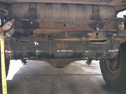 1984 Ford Bronco - The underside of a vehicle shows a rusty frame supporting a fuel tank. A yellow measuring tape is positioned to the left of the fuel tank,…