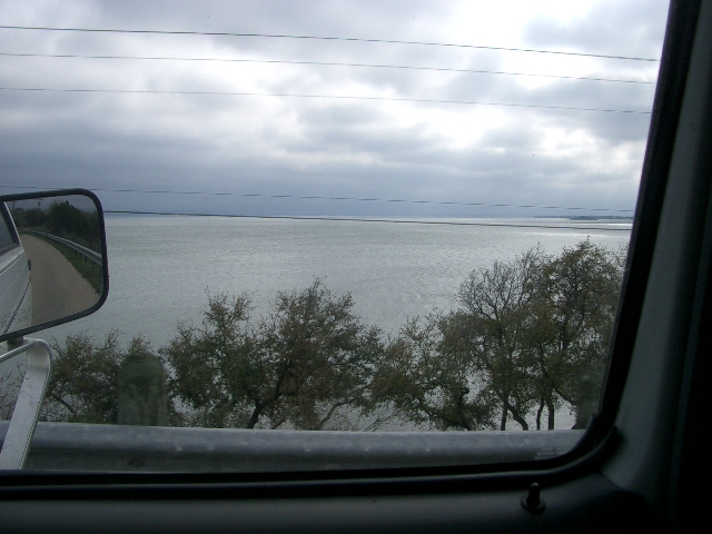 Side mirror view of a road leading to a large body of water under a cloudy sky