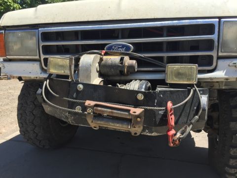 1984 Ford Bronco - The front bumper of a white Ford Bronco features a winch, tow hooks, and auxiliary lights. The vehicle is parked outdoors on a sunny day.
