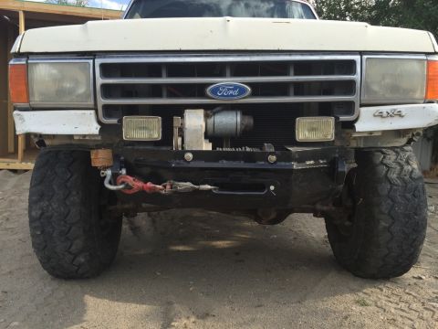 1984 Ford Bronco - Front view of a white Ford Bronco with an aftermarket black bumper, winch, and large off-road tires. The vehicle has a Ford emblem on the g…